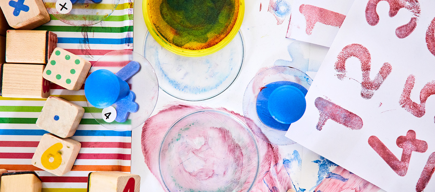 Bilder, die mit Wassermalfarben gemalt wurden, liegen auf einem Tisch. Daneben liegen bunte Würfel.